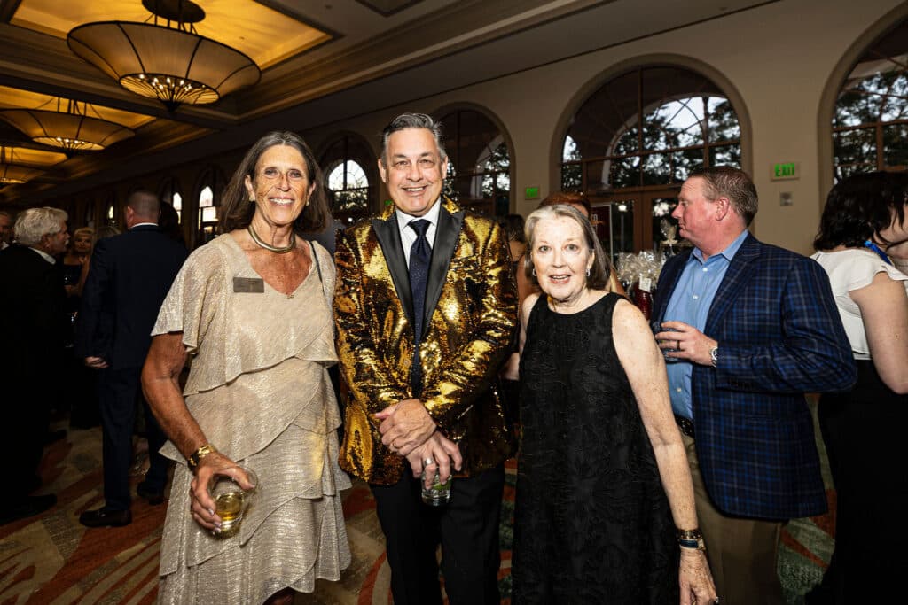 Three people pose for a photo at a formal event; a woman in a silver layered dress with a drink, a man in a shiny gold suit with a black shirt, and a woman in a black dress with floral patterns, all smiling, with other attendees in the background.