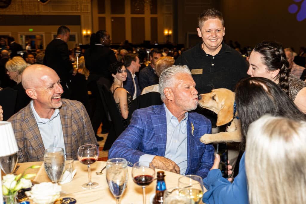 A lively gathering at a banquet where a man in a blue plaid suit is playfully kissing a golden retriever puppy. Nearby, a smiling woman in a black shirt watches, and a group of guests at tables is socializing in the background. The setting includes elegantly arranged tables with drinks and floral arrangements.
