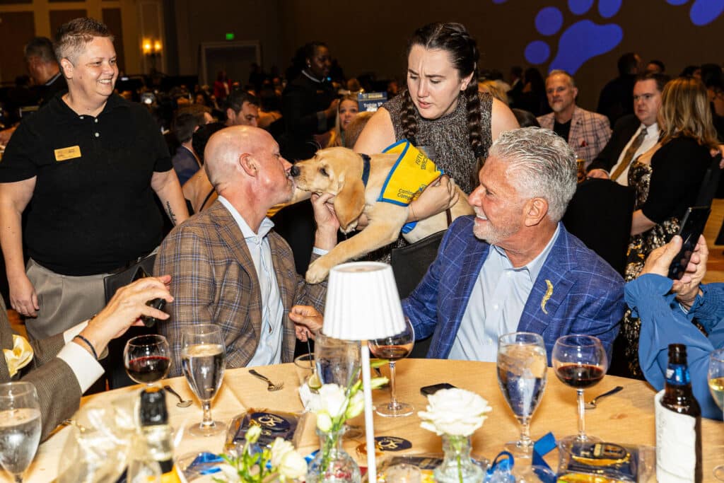 A lively event scene featuring a man kissing a golden retriever puppy wearing a blue and yellow service vest, while a woman holds the puppy. In the background, several guests are seated at tables with drinks and food, and a staff member is smiling nearby.