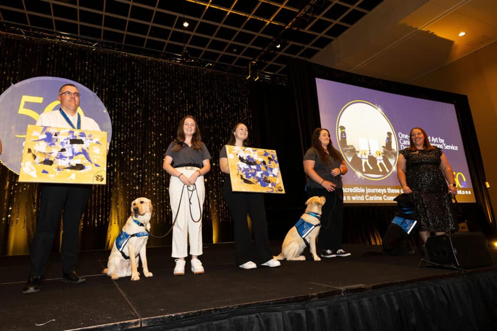 A group of four individuals stands on stage at an event, each holding a painting. Two Labrador retrievers sit beside them, wearing vests. The backdrop features a sparkling curtain and a screen displaying information related to the event.
