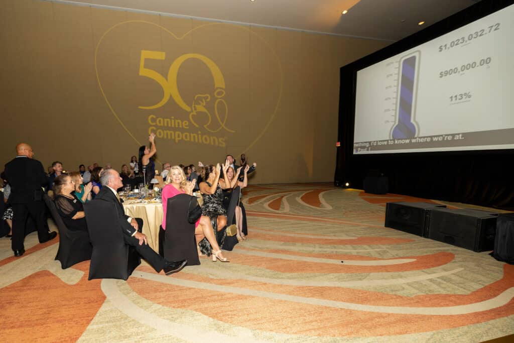 A lively event scene at a gala for Canine Companions, featuring attendees cheering and clapping, with a large screen displaying a fundraising thermometer reaching over $1 million, and the logo for the 50th anniversary projected on the wall behind them.