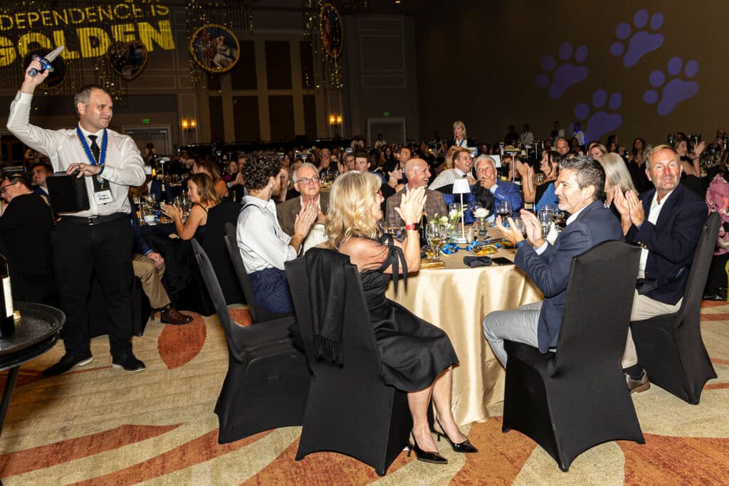 A banquet scene where a waiter is holding a bottle with a corkscrew, while guests at elegantly set tables applaud and celebrate. The room is decorated with festive elements, including a large banner above that reads 'Independence is Golden.' Many attendees are dressed formally, with some laughing and enjoying drinks.