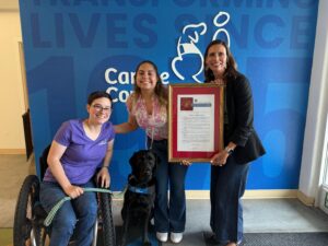 Three individuals pose together in an office setting with a blue wall featuring the words "TRANSFORMING LIVES SINCE". One person in a wheelchair holds a leash attached to a black service dog, while another person stands next to them holding a framed resolution. The third person stands on the other side, smiling at the camera.