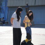 Two women are standing outdoors, one wearing a gray t-shirt and the other a yellow tank top, both looking down at a black dog wearing a yellow service vest. The dog is sitting on the pavement, and a statue is visible in the background.