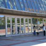 Two people walking by the entrance of the Edwin W. Pauley Pavilion, a modern building with large glass doors and reflections showing the outdoor environment; one person is walking a black dog while the other is checking their phone.