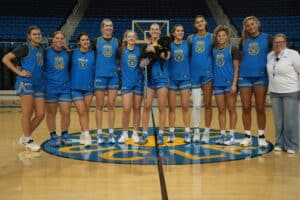 A group of twelve UCLA women's basketball players and coach Cori Close pose together on a basketball court. They are wearing blue UCLA jerseys and shorts, with various numbers displayed. One player is holding a black dog. The court features the UCLA logo at center and empty blue seats in the background.