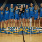 A group photo of the UCLA women's basketball team on the court, wearing blue and gold uniforms, with nine players smiling and posing together, while one player holds a small black dog. A coach or staff member stands off to the side, smiling at the group.
