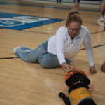 A woman wearing a UCLA-branded white sweatshirt and blue pants lies on the hardwood floor of a gym, reaching out to a black dog wearing a blue service vest and an orange basketball on its head, while another person is kneeling beside them.