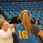 A woman with glasses smiles while playfully interacting with a black dog held by a person in a blue athletic jersey with the number 16. They are inside a sports arena with blue seats in the background.
