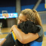 A young woman with long, wavy hair hugs a black puppy close to her face in a sports arena, with blue bleachers in the background. The puppy has a yellow vest on. Cheerleaders can be seen training in the background.