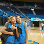 Two smiling female basketball players, wearing blue jerseys, pose together on a gym court while holding a black dog. The background shows empty bleachers and other players practicing.