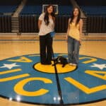 Two women standing on a basketball court with a black dog lying on the ground between them; the court features a prominently displayed logo with blue and yellow colors.
