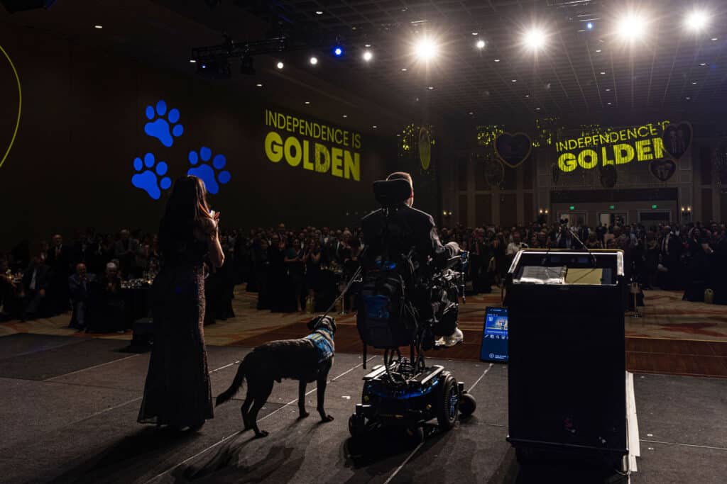 A woman in a sparkling gown stands on stage clapping, while a man in a wheelchair, facing away from the camera, listens. A black dog with a blue vest is beside him. The backdrop features large text that reads 'INDEPENDENCE IS GOLDEN' and blue paw prints, with a crowd of attendees in formal attire watching.