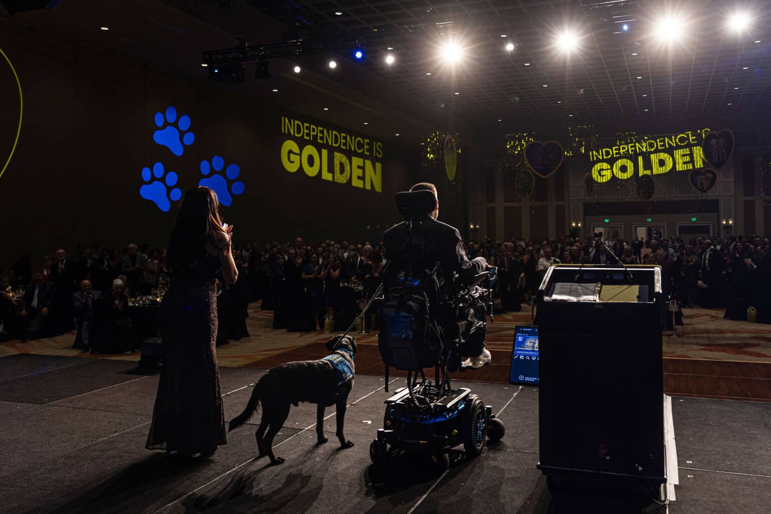 A woman in a sparkling gown stands on stage clapping, while a man in a wheelchair, facing away from the camera, listens. A black dog with a blue vest is beside him. The backdrop features large text that reads 'INDEPENDENCE IS GOLDEN' and blue paw prints, with a crowd of attendees in formal attire watching.