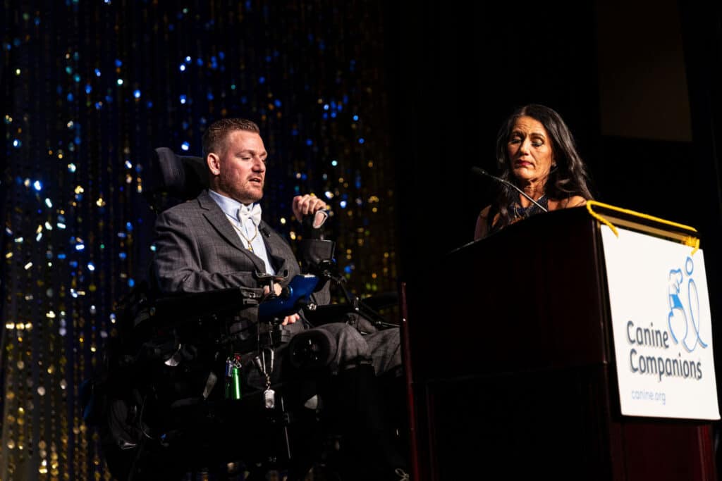 A man in a wheelchair wearing a suit and bow tie speaks at a podium, while a woman in a dark dress stands beside him. The background features golden sequins, adding to the event's formal atmosphere.