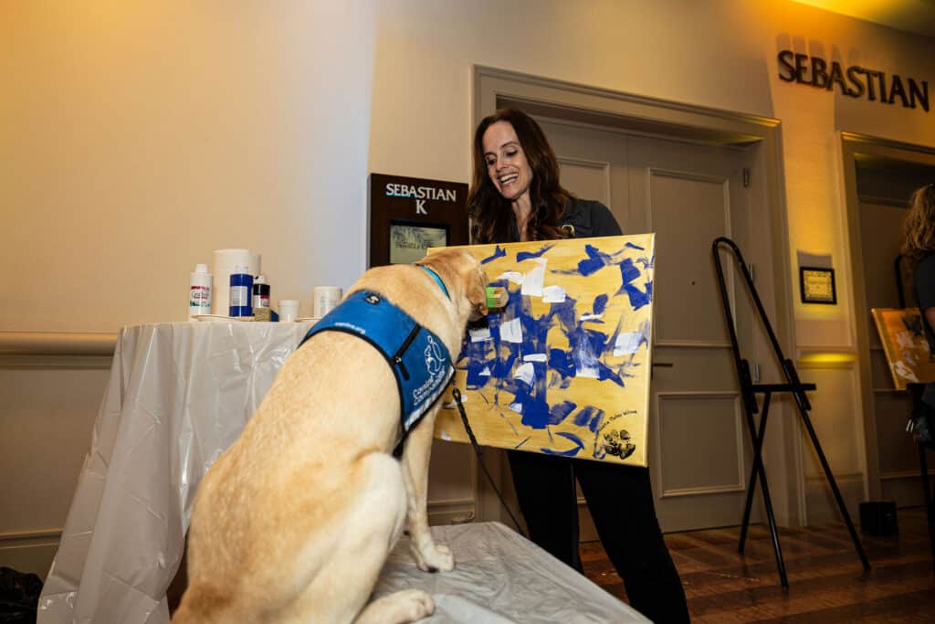 A woman smiles as she interacts with a yellow Labrador retriever that is painting on a canvas. The dog is wearing a blue harness and has its paw on the canvas which features blue and white paint. In the background, there are art supplies on a covered table and a sign that reads 'SEBASTIAN.' The setting appears to be an art event.