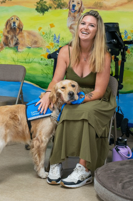 A woman in a green dress sits on a chair, smiling while gently petting a golden retriever wearing a blue therapy dog vest. The background features a colorful mural with images of dogs and greenery.