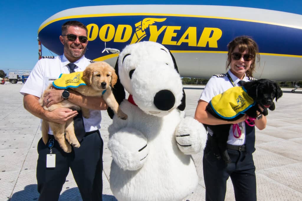 Two pilots, one male and one female, are posing with a large costumed character resembling Snoopy in front of a Goodyear blimp. The male pilot is holding a golden retriever puppy, while the female pilot is holding a black puppy, both wearing "Canine Companions" vests.