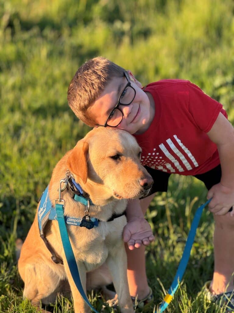 A young boy wearing glasses in a red t-shirt leans against a sitting yellow Labrador Retriever, smiling at the camera while in a grassy field during sunset.