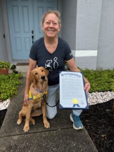 A smiling woman in a black t-shirt crouches next to a golden retriever puppy, holding a proclamation document, in front of a light blue door and landscaped yard.