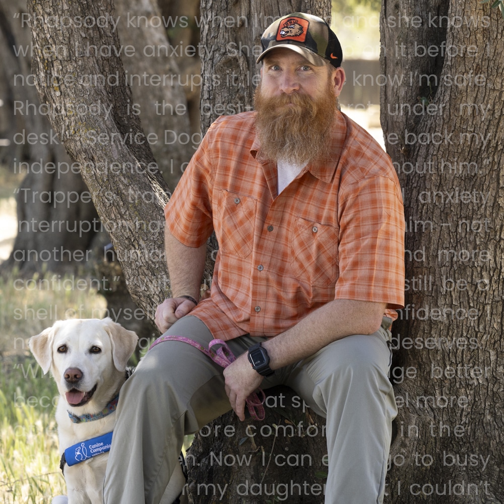 A man with a long beard wearing an orange plaid shirt and a baseball cap sits against a tree, beside a friendly yellow Labrador dog with a service dog vest. The man looks relaxed and is holding the dog's leash.