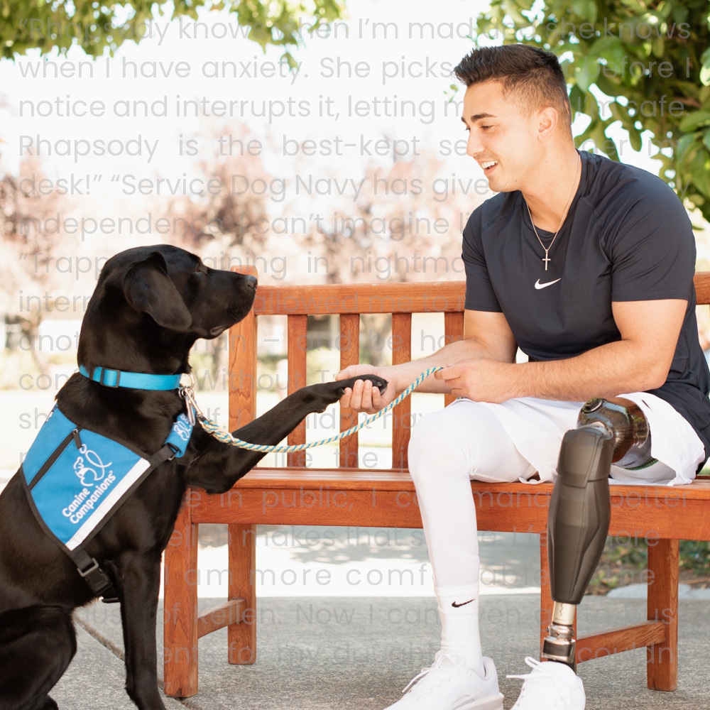 A man with a prosthetic leg sits on a bench, smiling as he holds paws with a black service dog wearing a blue vest. The setting is outdoors with trees in the background.