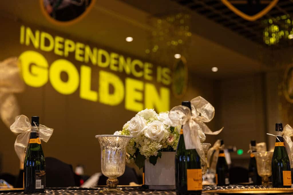 A table setting featuring champagne bottles adorned with ribbons, a floral centerpiece with white roses and greenery, and a glass vase. In the background, the words 'INDEPENDENCE IS GOLDEN' are projected in large, yellow letters on the wall.