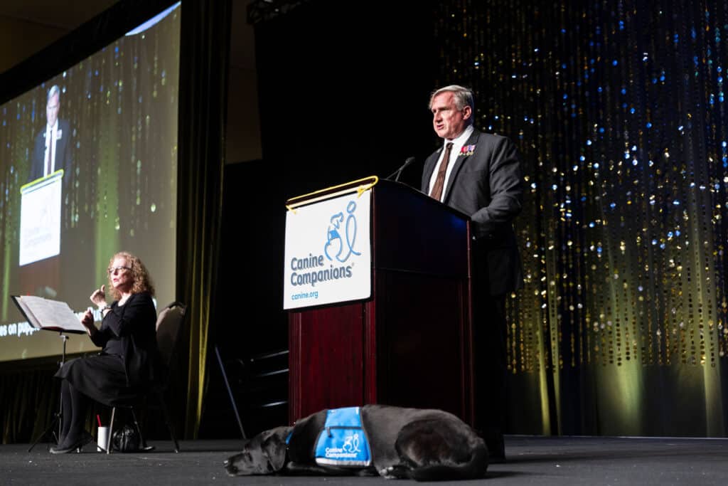 A man in a suit speaks at a podium bearing the Canine Companions logo, while a woman in a black dress sits beside him gesturing; a black dog with a blue service vest lies quietly on the stage.
