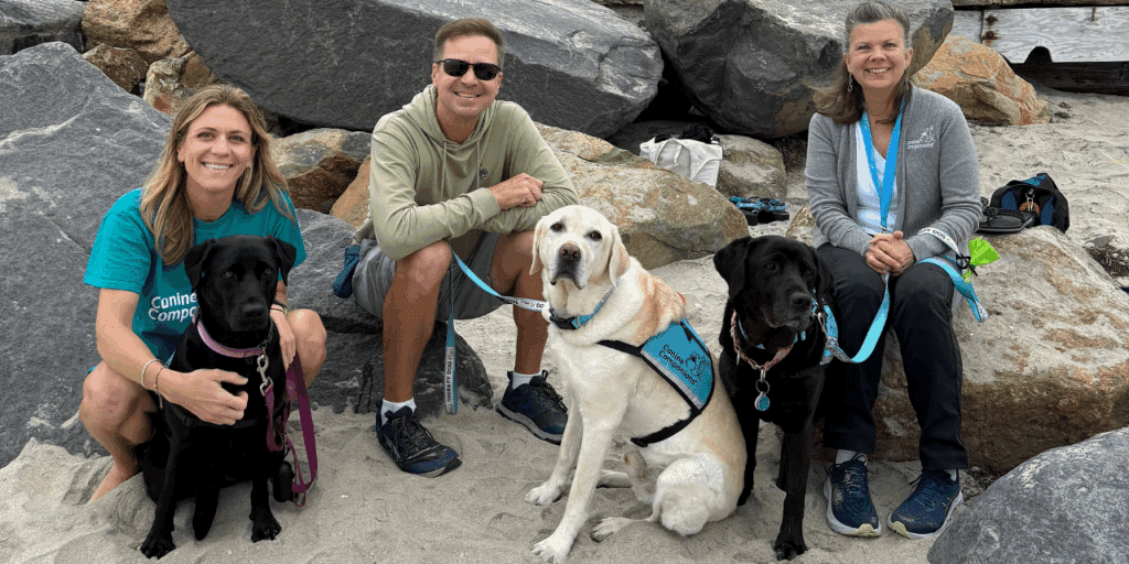 A group of four people and three dogs are sitting on a sandy beach near large rocks. A woman in a teal shirt smiles while holding a black Labrador Retriever on a leash. A man in a green sweatshirt poses with a yellow Labrador wearing a blue service vest. Next to him sits a woman in a gray sweater holding a black Labrador, and another black Labrador is resting on the sand.