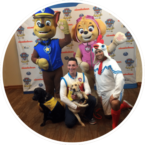 A man in a blue and white costume poses with a blonde dog while another man kneels beside them. They are flanked by two mascot characters: a brown dog in a police outfit and a yellow dog with pink accents. Two real dogs, one black and one light-colored, are both present in the scene. The background features a Nickelodeon promotional backdrop.
