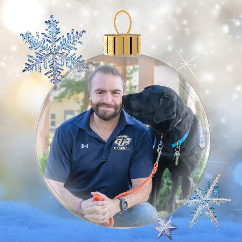 Bearded man in a navy baseball polo sits smiling as a black Labrador wearing a service-dog vest nuzzles his cheek, both pictured inside a glass Christmas ornament surrounded by sparkly snowflakes and a blue snowy foreground.