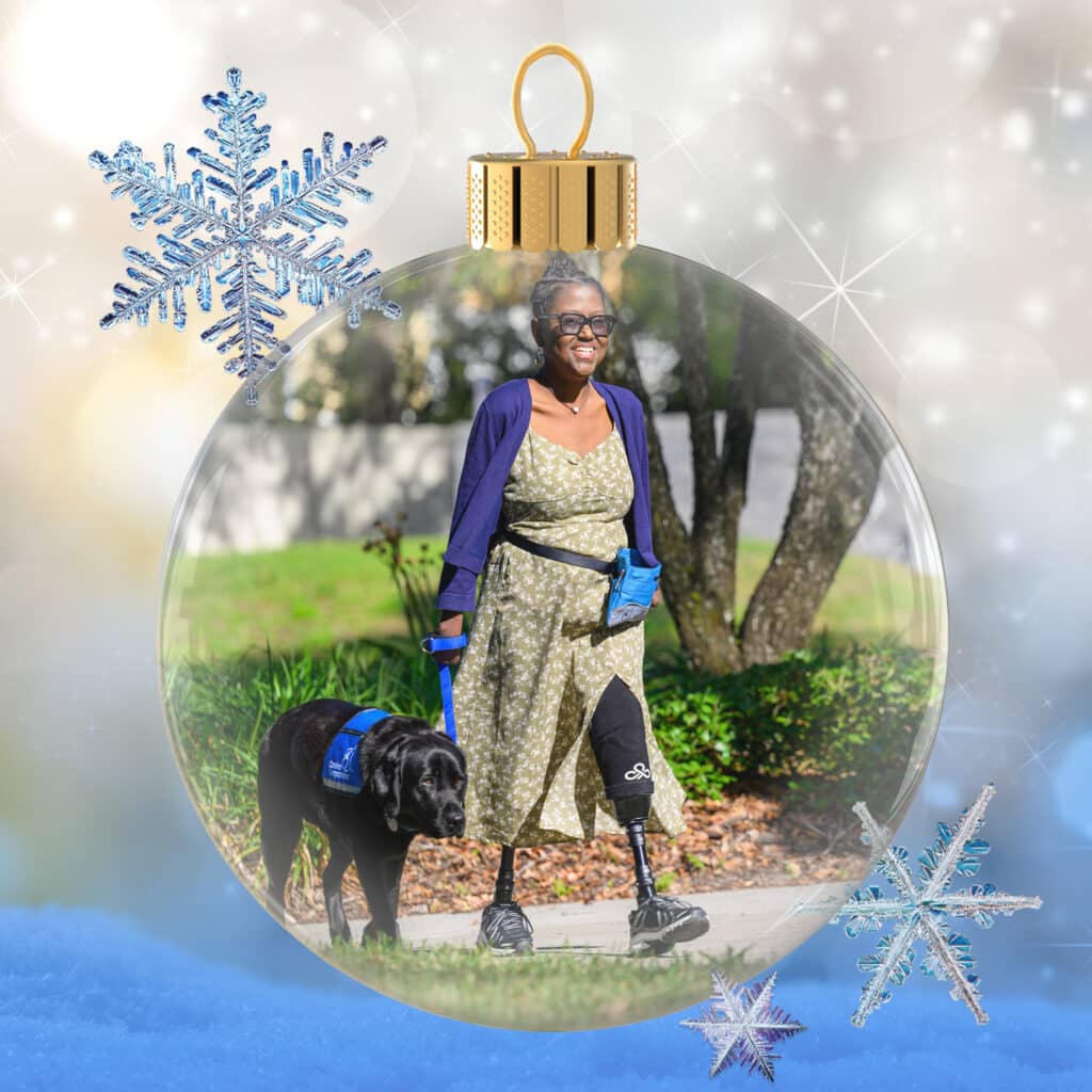 Smiling woman wearing sunglasses, a floral green dress and blue cardigan walking a black service dog while using two prosthetic legs, pictured inside a decorative glass Christmas ornament with snowflake accents and a sparkly bokeh background.