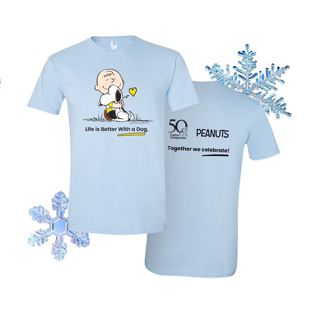 Two light-blue T-shirts shown front and back: the front displays Charlie Brown hugging Snoopy with a small yellow heart and the caption Life is Better With a Dog, and the back shows a 50 Canine Companions logo, the word PEANUTS, and the slogan Together we celebrate!; blue snowflake graphics surround the shirts.