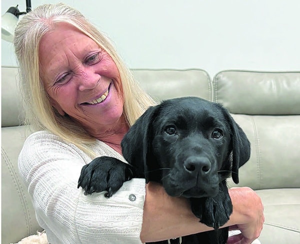 A smiling older woman with long blonde hair cradles a black Labrador puppy on a light-colored leather sofa.