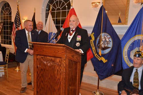 Elderly man in formal black suit with medals speaks at a wooden podium, flanked by military flags (including Navy and Air Force), while two men stand to his left and a seated veteran watches on the right.