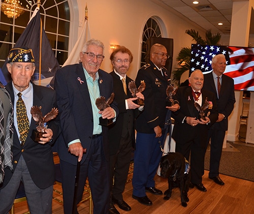 Group of men, some in military uniform or veterans’ caps, standing in a banquet room holding small eagle trophies with an American flag backdrop and a black service dog at their feet.