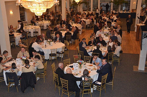 Overhead view of a large formal banquet in a ballroom with guests seated at round tables adorned with centerpieces, gold chairs, a crystal chandelier overhead and people mingling around the room.