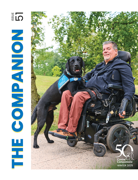 A man in a wheelchair smiles while sitting outdoors, accompanied by a black Labrador dog wearing a service vest. The background features greenery and a pathway, suggesting a pleasant outdoor setting.