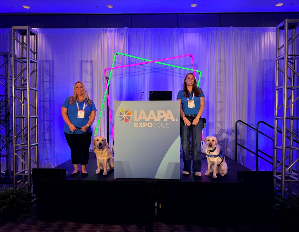 Two women in blue shirts stand on either side of a podium labeled IAAPA Expo 2025 on a stage with blue lighting and neon geometric light frames, each holding a leash attached to a seated yellow Labrador service dog wearing a vest.