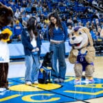 UCLA students and puppy raisers Audrey and Kyubin, along with black Labrador and future service dog Pauley, wearing a UCLA jersey and Canine Companions vest, stand at center court amid UCLA cheerleaders and two mascots—a brown bear and a tan dog—holding blue and gold pom-poms with a crowd in the arena behind them.