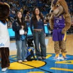 Two young women stand at center court with a black service dog sitting between them, flanked by UCLA mascots and cheerleaders holding blue and gold pom-poms in front of a crowded arena.