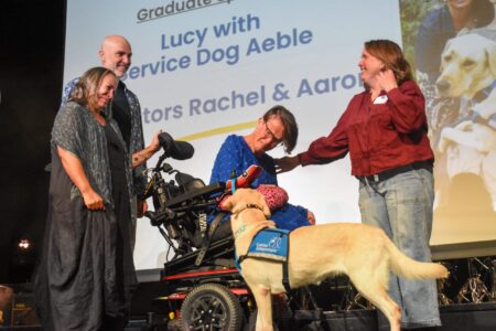 Woman in a wheelchair on stage leans forward to pet a yellow Labrador wearing a blue Canine Companions service vest while three smiling adults stand around and a presentation slide is projected behind them.