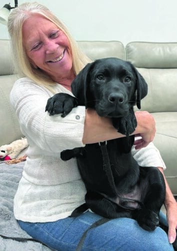 Smiling woman with long blonde hair sitting on a couch, cradling a black Labrador puppy in her lap as the puppy looks toward the camera with its paws draped over her arm.