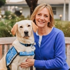 Smiling woman in a blue sweater sits on a bench with a yellow Labrador retriever wearing a blue service vest.