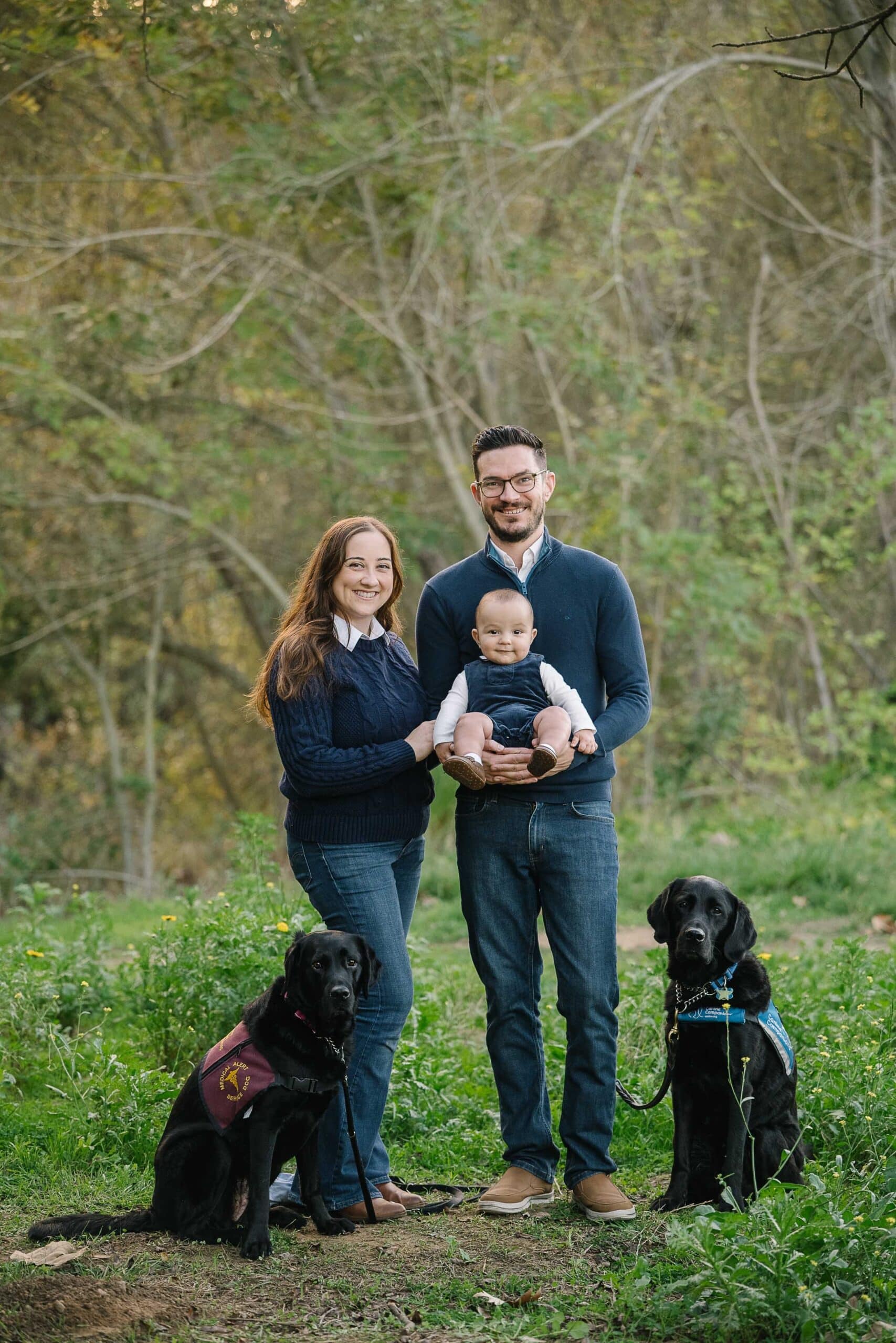 Smiling couple in navy sweaters with father holding their baby, flanked by two black lab service dogs wearing vests.