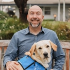 Smiling man seated on a bench with a yellow Labrador wearing a blue service vest.