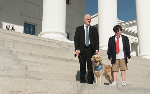 Bald man in a suit and a school-aged boy in a blazer and red tie standing on wide stone steps beside a golden retriever wearing a blue bandana.