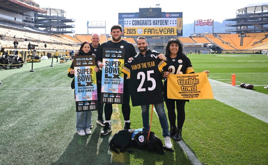 Five people in Pittsburgh Steelers apparel stand on the field at Acrisure Stadium holding oversized Super Bowl tickets, a 'Fan of the Year' jersey numbered 25 and a yellow Terrible Towel, with a service dog lying at their feet and a scoreboard behind them reading 'Congrats Hayden! 2025 Fan of the Year'.