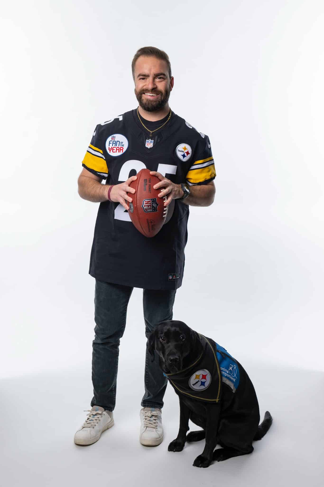 A smiling man in a black and yellow Pittsburgh Steelers jersey holds an NFL football while a black Labrador wearing a blue service-dog vest with a Steelers patch sits at his feet against a plain white background.