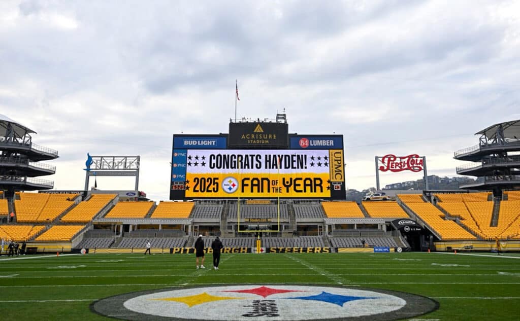 Acrisure Stadium's empty Pittsburgh Steelers field with the team's logo painted at midfield and a large central scoreboard reading 'Congrats Hayden! 2025 Fan of the Year' above rows of yellow seats.